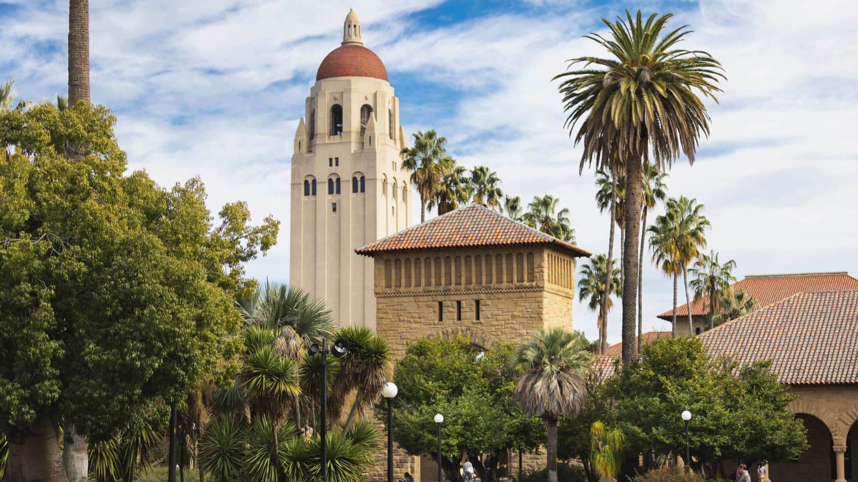 A scenic view of Stanford University featuring the iconic Hoover Tower in the background, surrounded by palm trees and lush greenery. In the foreground, students are sitting on a circular bench and walking along the paths, engaging with their devices and studying. The clouds in the blue sky create a picturesque atmosphere over the campus.