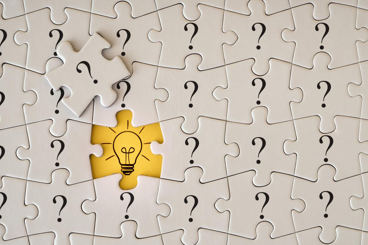 A close-up image of a puzzle featuring predominantly white pieces with black question marks on them. In the center, there is a missing piece where a yellow background reveals a light bulb symbol, suggesting an idea or solution. A single puzzle piece rests beside the incomplete area, also decorated with a question mark.