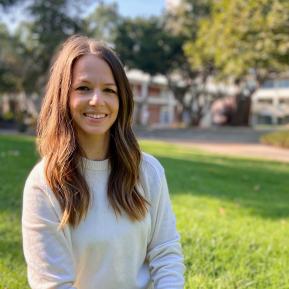 Image of female wearing a white sweater, standing with grass in the background and a building in the distance.