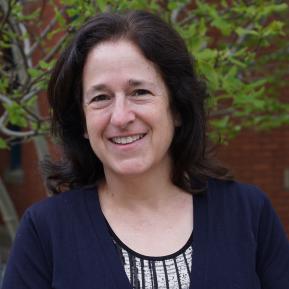 Image of female with black hair till their shoulders, wearing a blue cardigan standing in front of a tree.