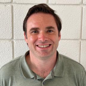 Image of white male wearing an olive green shirts standing in front of a white brick background