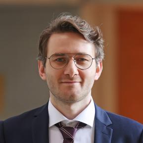 image of male with glasses and brown hair wearing a blue suit with a white shirt