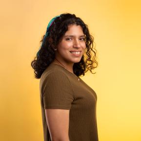 image of female with curly hair wearing a yellow top standing in front of a yellow background