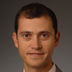 A person wearing a light pink shirt and a gray suit with a striped tie poses against a dark background.