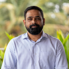 Man with short dark hair and beard, wearing a light checkered button-up shirt, standing outdoors with blurred green foliage in the background.