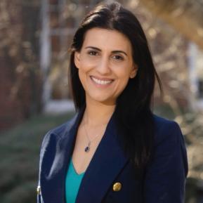 A person wearing a dark blue blazer over a green top stands outdoors, with a blurred background featuring trees and buildings.