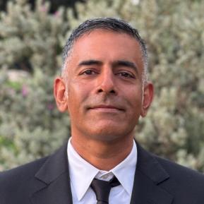 The individual is dressed in a formal black suit with a white dress shirt and a black tie. They stand in front of a backdrop filled with greenery, creating an elegant and composed atmosphere. The lush foliage adds a touch of nature to the formal attire, enhancing the overall presentation.