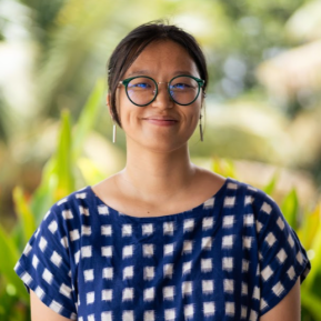 Smiling woman with glasses and tied-back hair, wearing a blue patterned top, standing outdoors with green foliage in the background.