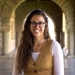 A person with long, wavy hair wearing a light-colored long-sleeve shirt under a brown vest stands in a corridor adorned with arches and soft lighting.