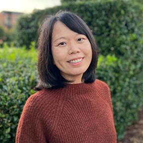 A person stands in a lush green outdoor setting, wearing a cozy, textured rust-colored sweater. The background features well-manicured hedges, creating a serene and inviting atmosphere. The soft, natural lighting enhances the warmth of the scene.