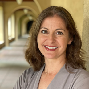 A person with wavy brown hair wearing a light gray top stands in a corridor with arches and soft lighting.