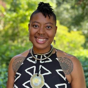 A woman stands outdoors in a green setting, wearing a black sleeveless dress adorned with white geometric patterns. She accessorizes with multiple necklaces, including a statement piece with a circular pendant. Intricate tattoos peek out from her arms, adding to her unique style and character. The background features soft greenery, creating a relaxed and natural atmosphere.