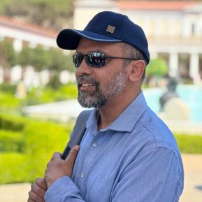 A person is standing outdoors in a scenic location, wearing a navy blue cap and a light blue button-up shirt. They hold a bag over their shoulder while appearing to enjoy the surroundings. In the background, there are well-maintained gardens and an elegant building, suggesting a picturesque and tranquil setting ideal for a leisurely visit. The sunlight illuminates the scene, enhancing the vibrant greenery and architecture.