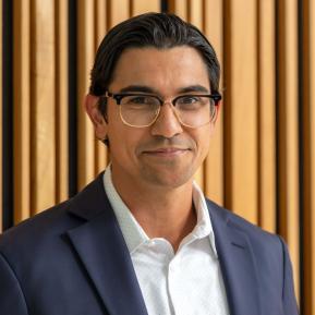 Individual dressed in a dark blazer over a light-colored shirt with a subtle polka dot pattern. The setting includes wooden paneling in the background, adding a modern and sophisticated vibe to the overall scene. The individual appears confident and approachable, typical of a professional portrait. Additionally, the styling suggests a contemporary and polished look suitable for formal or business environments.