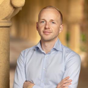 A man stands confidently with his arms crossed, wearing a light blue button-up shirt. The background features warm-toned architectural elements, suggesting an outdoor or semi-outdoor setting with soft natural lighting. The environment reflects an inviting and professional atmosphere, enhancing the overall composure of the individual.