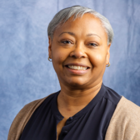 A close-up portrait of a woman with short, gray hair, wearing a dark blouse and a light brown cardigan. The background is a soft blue gradient. The woman is smiling and facing slightly to the side, exuding a professional and approachable demeanor.