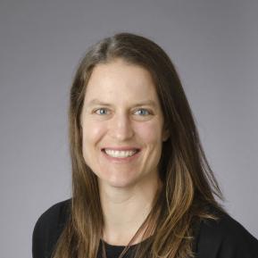 A woman with long, wavy brown hair is pictured in a professional setting. She is wearing a black top, which gives her a polished appearance. The background is a gradient of gray, contributing to the formal look of the portrait. The composition focuses on her shoulders and upper body, ideal for a professional profile or biography.