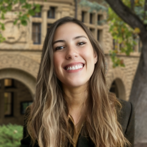 A woman stands outdoors in front of an architectural structure characterized by warm tones and intricate details. She has long wavy hair with highlights, and she is dressed in a dark blazer. The background features hints of greenery, along with elements of the building, creating a sophisticated and academic atmosphere. The image captures a moment of confidence and poise against a picturesque setting.