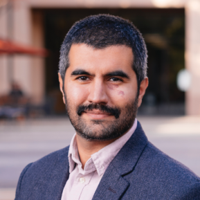 A person stands outdoors, wearing a navy blazer over a light-colored shirt. The background features a blurred urban setting with hints of outdoor seating and shaded areas, indicating a lively atmosphere. The image captures a moment of confidence and professionalism against a backdrop that suggests a casual yet vibrant environment.