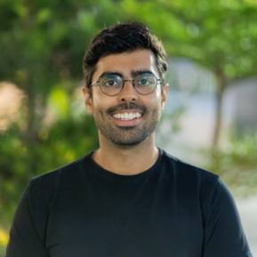 A person wearing a black t-shirt stands outdoors with a blurred background of greenery and softly illuminated lights. The setting appears to be during twilight, with a gentle warm ambiance created by the fading light.