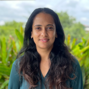 A person stands outdoors, wearing a flowing green blouse with long sleeves. The background showcases lush greenery and soft natural light, creating a serene atmosphere. The setting appears to be in a garden or park, with trees visible in the distance under a partly cloudy sky.