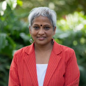 Smiling woman with short gray hair, wearing a red striped blazer and white top, standing outdoors in front of green foliage."