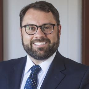 Individual wearing a dark suit jacket with a white shirt underneath and a patterned tie featuring blue with yellow polka dots. The background appears to include a well-lit indoor setting with subtle decorative elements, likely giving a professional or formal ambiance. The person has a neatly groomed beard, and the overall posture suggests a confident demeanor, typical in a portrait setting.