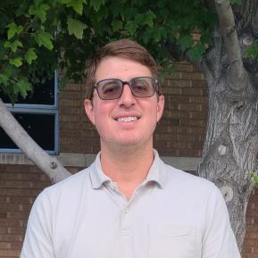 A person is standing outdoors in front of a brick wall and a tree. They are wearing a light gray polo shirt with a chest pocket. The setting is shaded by the tree, creating a relaxed atmosphere. The background features a window framed by the brick structure.