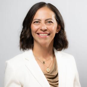 The subject is dressed in a tailored white blazer over a draped beige top. They are adorned with layered necklaces, one featuring a circular pendant and another with a decorative charm. The background is a soft gray, providing a neutral and elegant backdrop to the formal attire. The overall appearance conveys professionalism and sophistication.