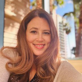 A person is sitting in a relaxed outdoor setting, dressed in a black top with a polka dot pattern and a light beige cardigan. Long, wavy hair cascades past the shoulders. The background features palm trees and a clear blue sky, creating a vibrant and sunny atmosphere. The warm light enhances the overall feeling of leisure and comfort.