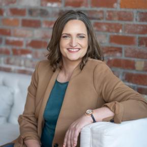 Seated in front of a textured brick wall, the subject is wearing a brown blazer over a teal top. They have a relaxed posture, with one arm resting on a white couch. A watch is visible on their wrist, adding a touch of elegance to the casual yet sophisticated outfit. The setting has a warm and inviting atmosphere, enhanced by the natural light.