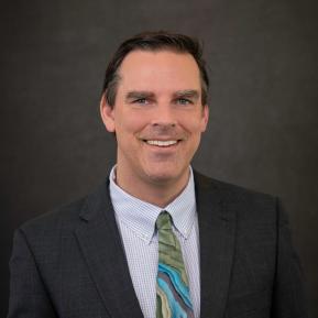 A man wearing a dark suit jacket and a checkered dress shirt with a colorful, patterned tie stands against a neutral gray background. The focus is on his professional attire, suggesting a formal occasion or business environment.