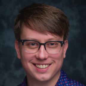 A person is dressed in a blue patterned shirt featuring red and white designs. The background is a dark textured surface, adding depth to the image. The overall impression is casual and relaxed, suitable for a friendly or informal setting.