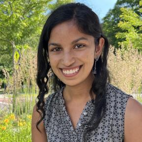 A person is wearing a sleeveless black and white patterned top, adorned with earrings, and is positioned outdoors among plants and flowers. The background features lush greenery and bright floral accents, contributing to a vibrant and natural setting.