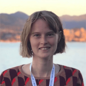 A person stands against a scenic backdrop of mountains and water during what appears to be sunset, with soft golden light illuminating the scene. They are wearing a multicolored patterned sweater and a lanyard around their neck. The atmosphere is serene and captures the beauty of a natural landscape.