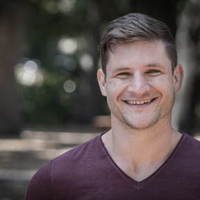 A young man stands outdoors in a natural setting. He is wearing a deep v-neck maroon t-shirt. The background features soft-focus greenery and blurred lights, creating a serene atmosphere. The lighting is bright, enhancing the fresh and vibrant look of the scene.