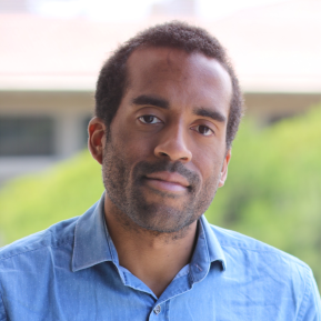 A person wearing a light blue, button-up shirt stands outdoors, with blurred greenery and a building visible in the background. The image is taken in daylight, creating a soft focus effect. The individual appears to be in a casual setting.