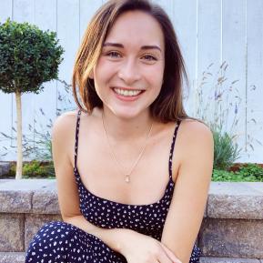 A person is sitting on a stone wall in an outdoor garden setting. They are wearing a dark dress with a floral pattern and a silver necklace. In the background, there is a white wooden fence and a small green shrub, along with some plants. The lighting is soft, indicating a pleasant outdoor environment.