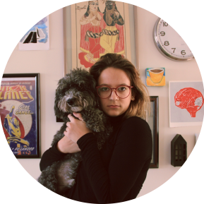 A person is holding a furry, black dog close to their chest. The background features a wall adorned with various framed artworks and a clock. The setting has a cozy and artistic ambiance, characterized by colorful prints and eclectic decor elements. The image is presented in a circular frame, enhancing its visual appeal.