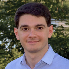 Headshot of a young man with short dark hair wearing a light blue button-down shirt, smiling slightly, with trees and greenery blurred in the background.