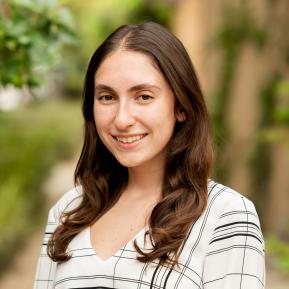 A portrait of a person with long, wavy brown hair wearing a white blouse with black stripes, with their face obscured. The background features greenery and a blurred pathway, suggesting an outdoor setting.