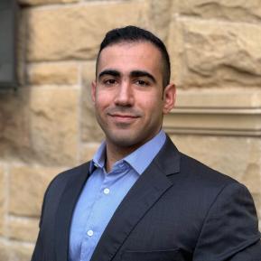A portrait of a person in a suit and dress shirt, with their face obscured. The background features a textured stone wall, indicating an outdoor or architectural setting.