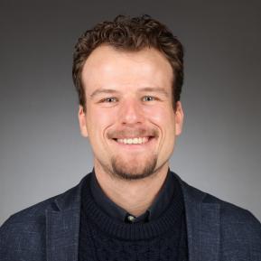 A portrait of a person with short, curly hair wearing a navy blue sweater and a jacket, with their face obscured. The background is a dark gradient, emphasizing the subject in the foreground.