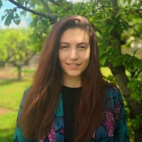 A portrait of a person with long, straight brown hair wearing a colorful patterned jacket over a black top, with their face obscured. The background features green foliage and a tree, indicating an outdoor setting.