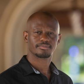 A portrait of a person with a shaved head wearing a black collared shirt, with their face obscured. The background features arches and soft, out-of-focus greenery, suggesting an outdoor or indoor setting with natural light.