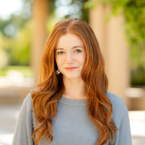 A portrait of a person with long, wavy red hair wearing a light blue sweater, with their face obscured. The background features soft greenery and architectural elements, indicating an outdoor environment.
