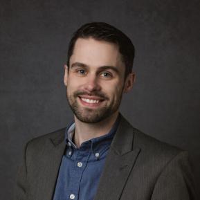 A portrait of a person wearing a dark blazer over a blue button-up shirt. The person's face is obscured with a blurred rectangle. The background is a dark, textured surface, giving the photo a professional look.