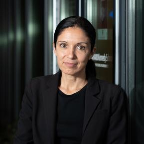 A person is seen wearing a black shirt and blazer, positioned against a reflective surface in a dimly lit environment. The background features muted lighting and vertical structures.