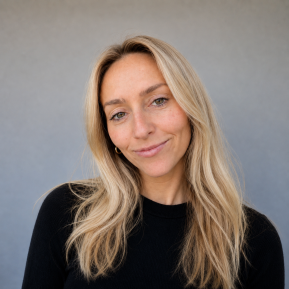 A portrait of a person with long, blonde hair wearing a black sweater. The person's face is obscured with a blurred rectangle. The background features a smooth, gray surface, enhancing the simplicity of the image.
