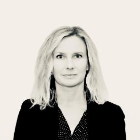 A person is depicted with shoulder-length blonde hair, wearing a black blazer over a white shirt with a polka dot pattern. The background is light-colored, and the image is presented in a stylized black and white format.